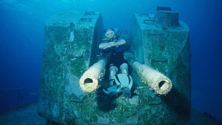 picture of diver underwater sitting on top of large gun from sunken ship